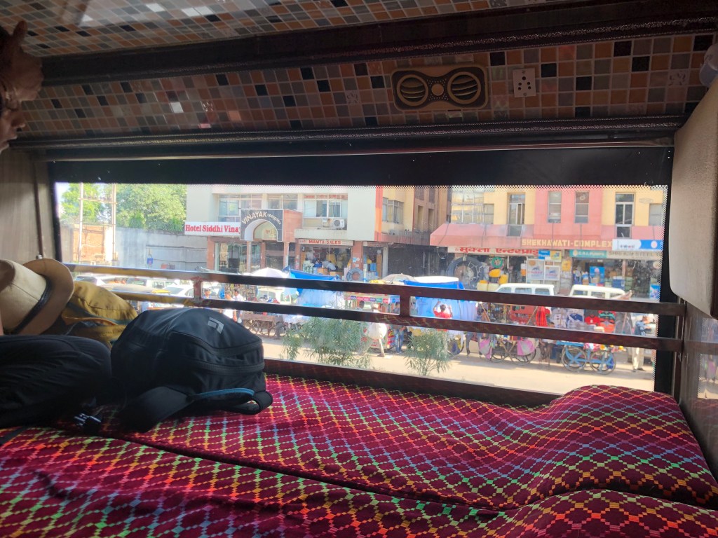 The cramped sleeper bus compartment. The hard and flat cushions are red with a rainbow checkered pattern and the window looking out onto an Indian street is wide and runs the length of the small cabin.