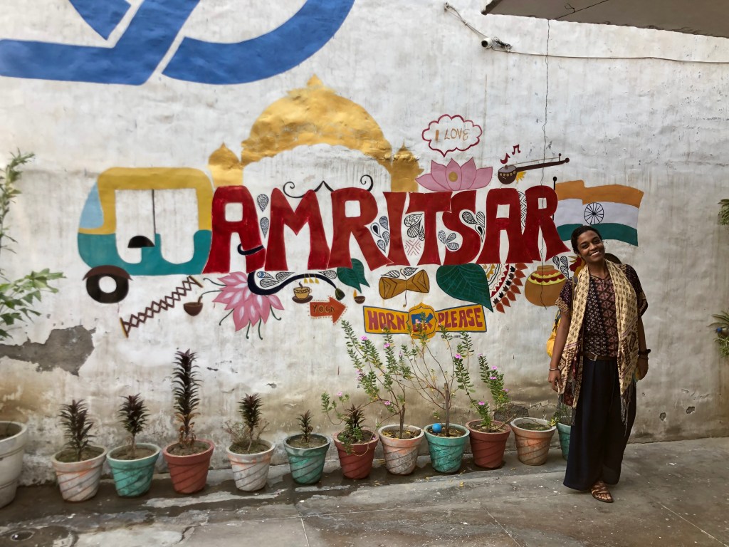 Amanda wears a scarf and boho style clothing while standing to the right of an Amritsar-themed mural.